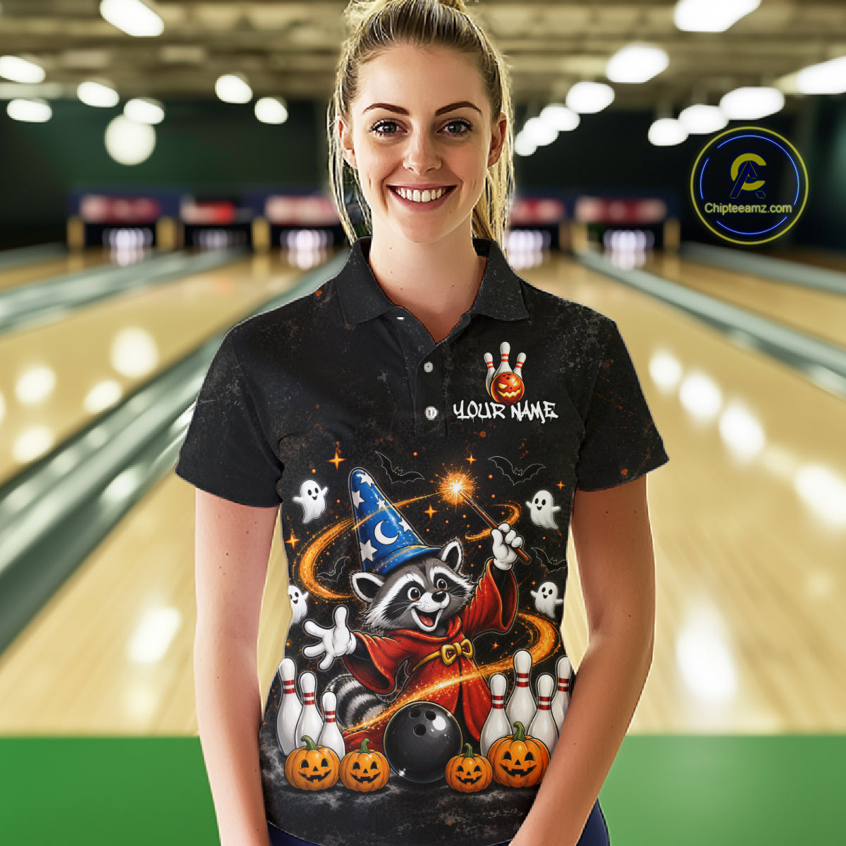 A woman stands in a bowling alley wearing a black short-sleeve polo shirt with a large Halloween raccoon graphic on the front. The shirt features a wizard hat, orange pumpkins, ghosts, and white bowling pins, with 'YOUR NAME' printed on the chest.
