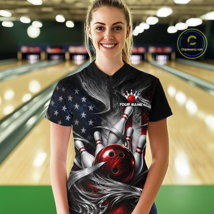 A woman stands in a bowling alley wearing a black short-sleeve bowling t-shirt with a custom graphic print featuring a red bowling ball, pins, and an American flag pattern. The shirt has a mock collar and 'YOUR NAME' printed on the chest.
