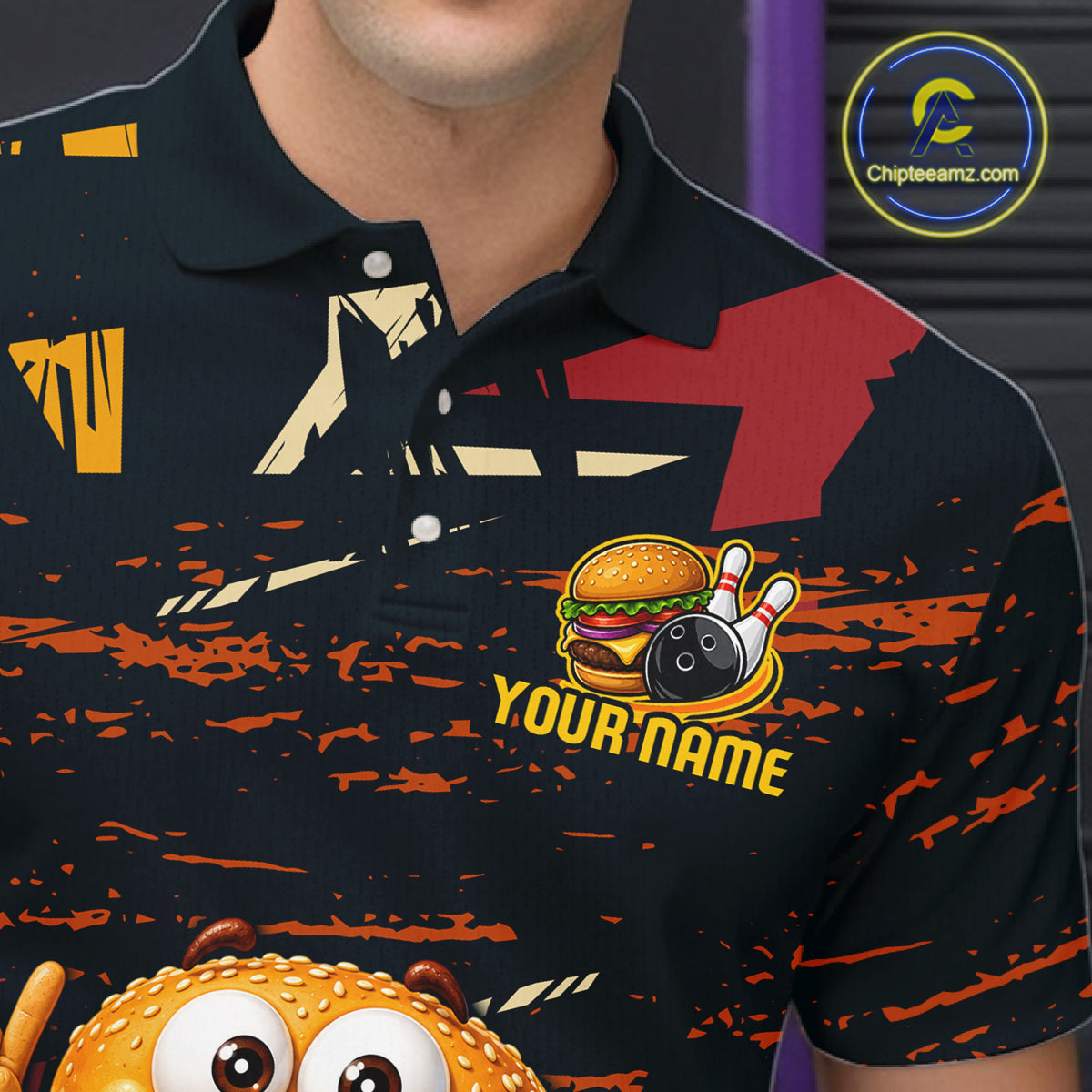 A close-up of a man wearing a black short-sleeve polo shirt with a colorful graphic design. The shirt features a red and beige abstract pattern, orange paint splatters, a cartoon burger with bowling pins and a bowling ball, and the text 'YOUR NAME' in yellow.
