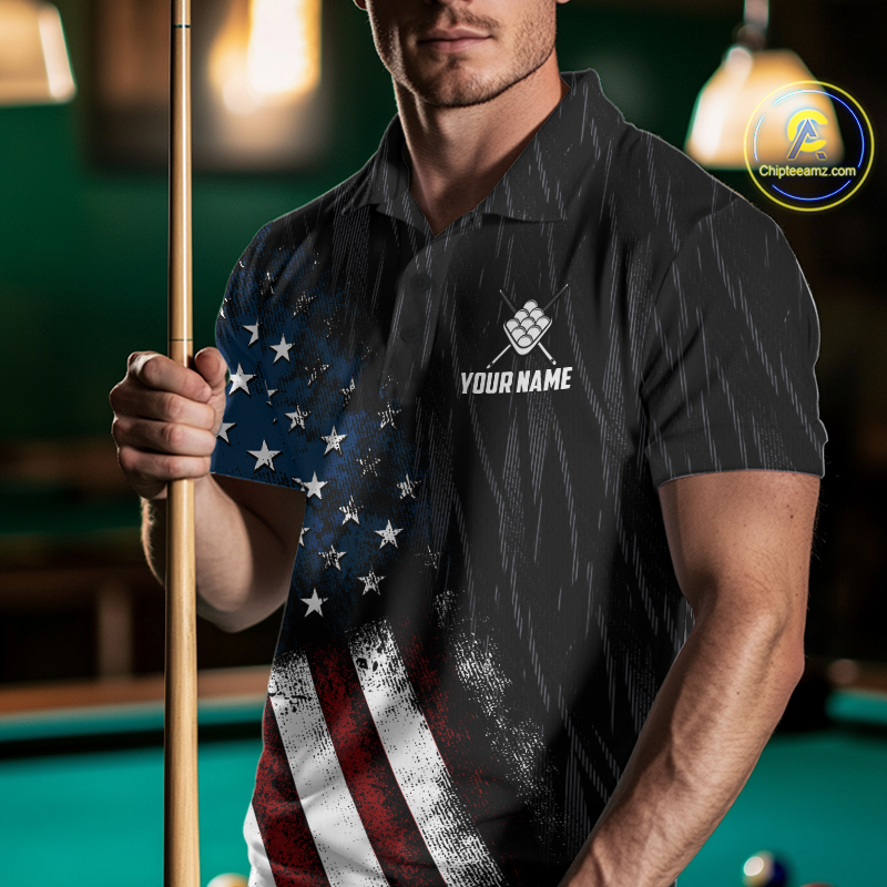 A man in a poolroom wearing a black polo shirt with a distressed American flag print on the right side and a white customizable logo on the chest. He is holding a pool cue, with a green pool table and warm hanging lights in the background.