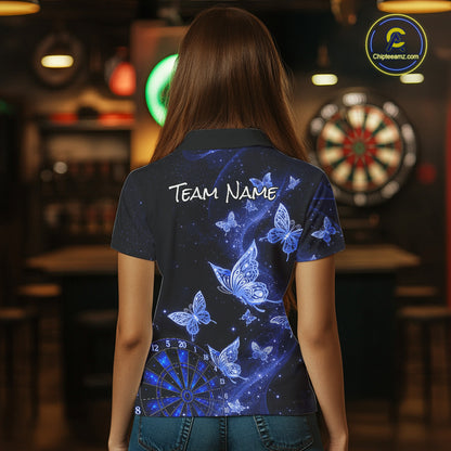 A woman viewed from behind wearing a dark navy short-sleeve polo shirt with a blue butterfly and dartboard print on the back, featuring the text 'TEAM NAME' in white. The background is a dimly lit bar with a dartboard visible.