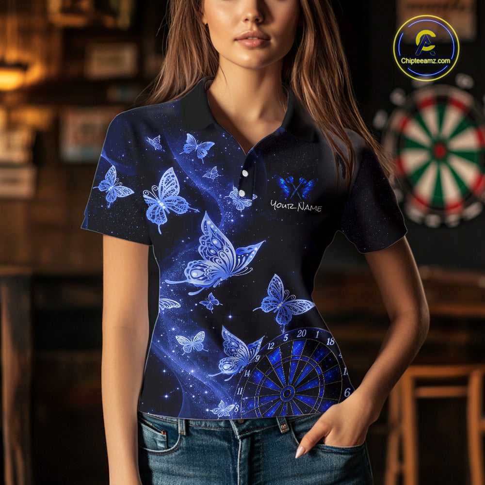 A woman wearing a short-sleeve polo shirt with a dark blue and black galaxy print, decorated with blue butterflies, stars, and a dartboard graphic. The shirt has a custom name printed on the chest, and the background shows a blurred indoor setting with a dartboard.