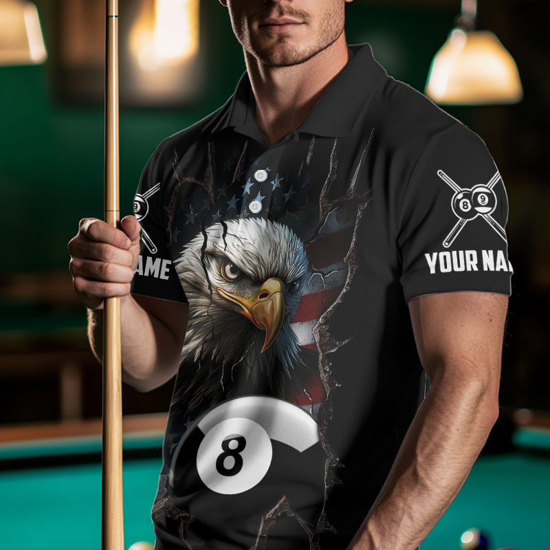 Man wearing a black polo shirt with an eagle and pool ball design, holding a pool cue in a billiards setting.