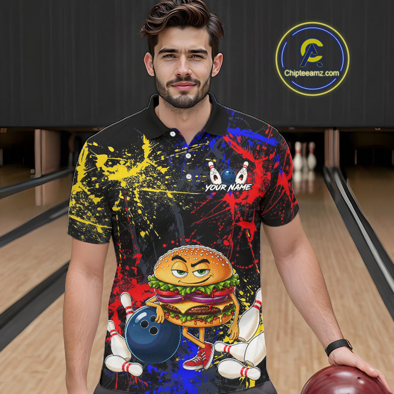 A man in a bowling alley wearing a black short-sleeve polo shirt with a colorful graphic print featuring a cartoon burger, bowling pins, bowling balls, and paint splatters. He is holding a bowling ball, with a circular 'ChipteeAmz'.