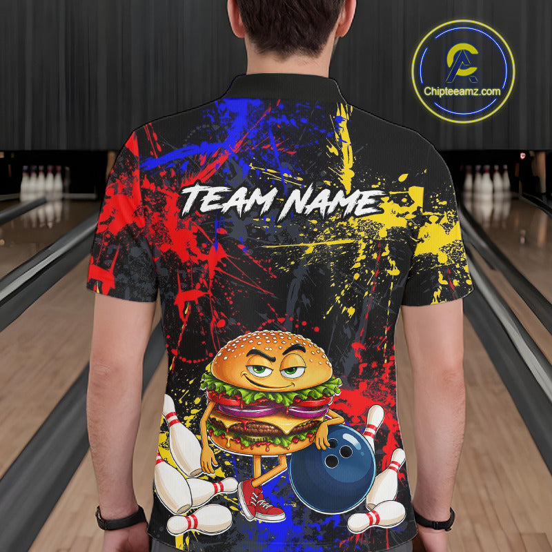 A man viewed from behind wearing a black short-sleeve bowling-themed t-shirt with a colorful red, blue, and yellow paint splatter design, a cartoon burger character holding bowling pins and a bowling ball, and the text 'TEAM NAME'. The background shows bowling lanes.