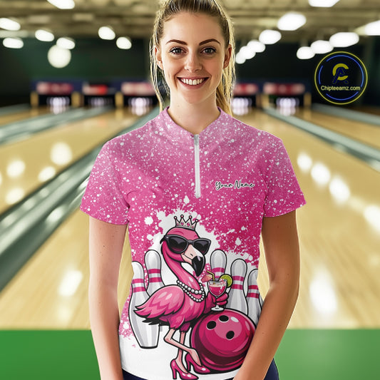 Female model at a bowling lane wearing a pink bowling quarter-zip shirt with a crowned flamingo, cocktail, bowling ball, and paint splatter graphics.