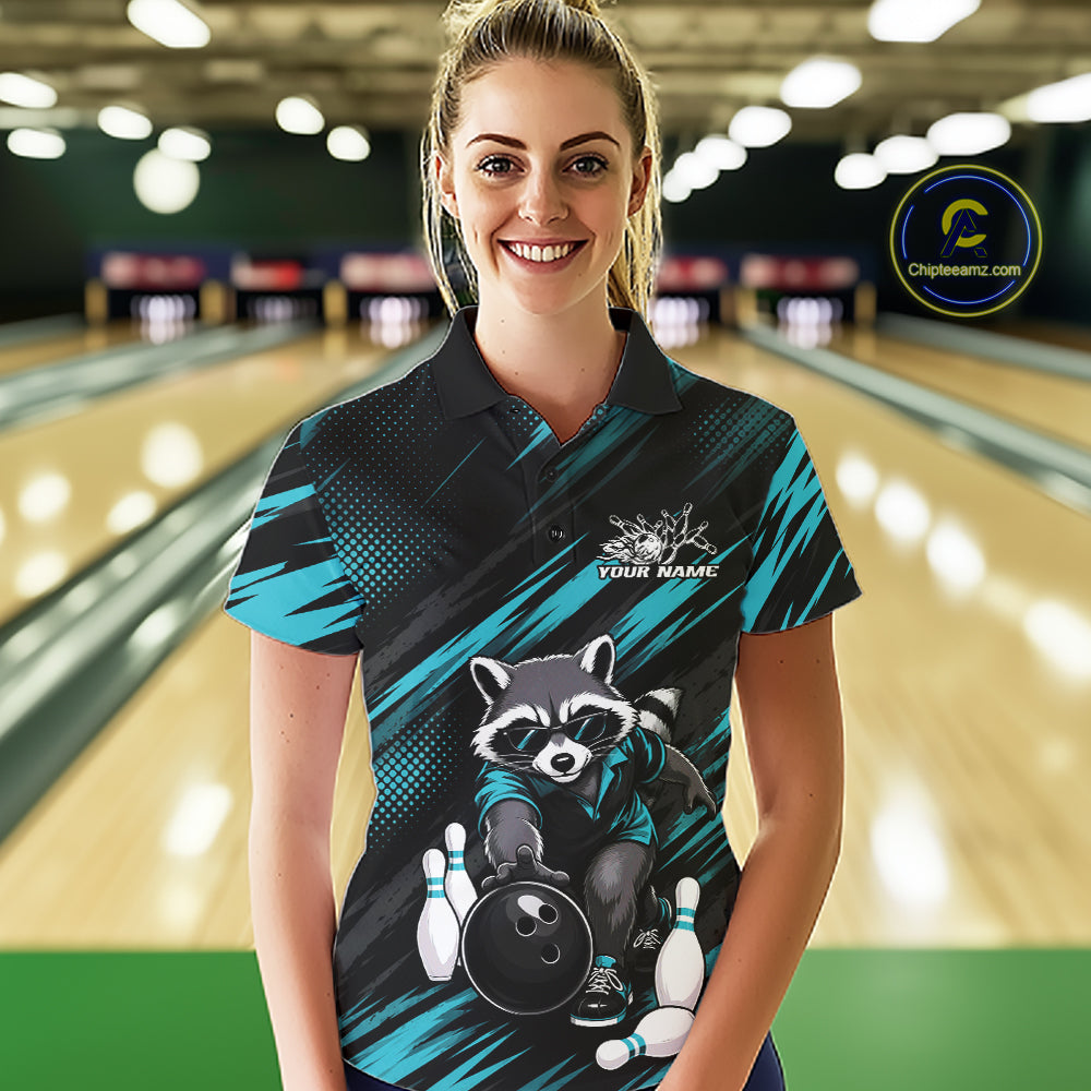 A woman stands in a bowling alley wearing a short-sleeve polo shirt with a raccoon graphic and teal diagonal stripes. 
