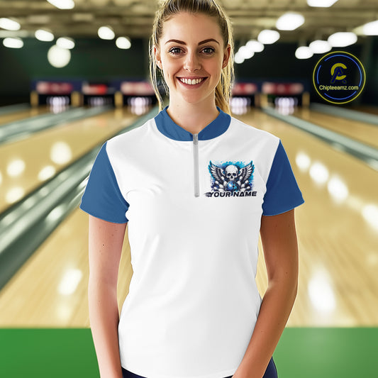 A woman stands in a bowling alley wearing a bowling shirt with quarter zip collar, blue skull-themed bowling graphic with black wings, pins, bowling ball