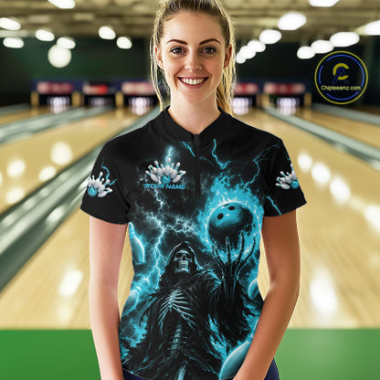A woman stands in a bowling alley wearing a black short-sleeve polo shirt with a dramatic blue and black graphic print featuring lightning, a skull, and a bowling ball. The shirt has a mock collar and sleeve buttons.