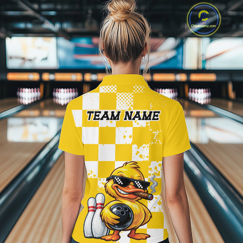 A woman viewed from behind wearing a bright yellow bowling-themed polo shirt with a checkerboard pattern, a cartoon duck graphic, and the text 'TEAM NAME' on the back. The background shows blurred bowling alley lanes and pins.