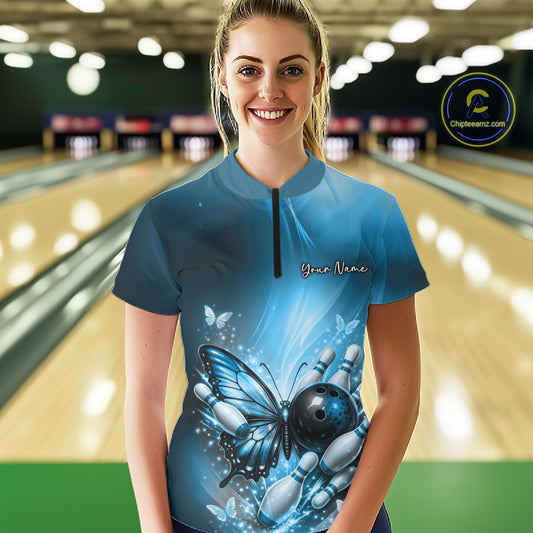 Female model wearing a blue bowling quarter-zip shirt at a bowling lane with butterfly artwork, a bowling ball, and scattered pin graphics.
