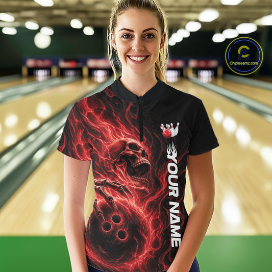 Front view of a female bowler in a bowling lane wearing a black bowling quarter-zip shirt with a swirling red flame skull gripping a bowling ball
