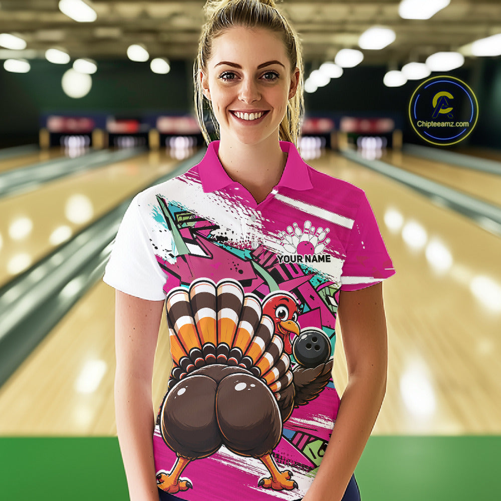 Front view of a female model wearing a pink custom bowling polo shirt with a funny graffiti turkey holding a bowling ball, paint splatter accents, and a personalized name in a bowling alley.