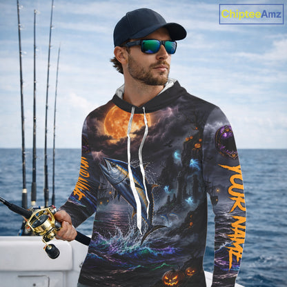 A man wearing a black hoodie with a detailed all-over Halloween fishing graphic print stands on a boat deck. The hoodie features a large blue fish jumping from the water under a glowing orange full moon, with jack-o'-lanterns and orange vertical text on the sleeves.