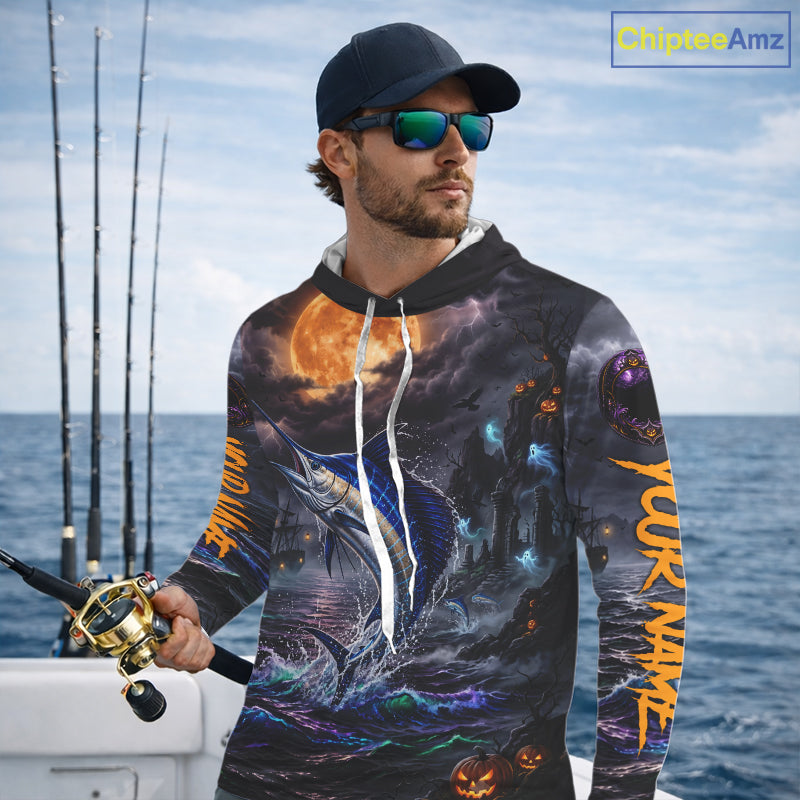 A man wearing a black baseball cap and sunglasses stands on a boat holding a fishing rod, wearing a custom all-over print hoodie featuring a blue marlin, a full moon, and orange text on the sleeves.