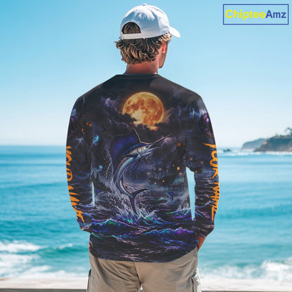 A man stands with his back to the camera wearing a long-sleeve graphic shirt featuring a dramatic print of a marlin jumping from the ocean under a large full moon, with blue and orange tones. The background shows a sunny beach scene with the ocean and sky.