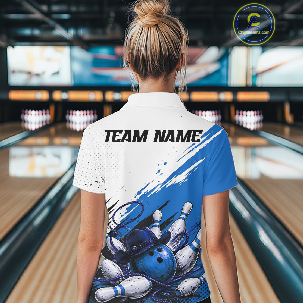 A woman viewed from behind wearing a custom bowling-themed short-sleeve polo shirt with a white and blue design featuring bowling pins and a blue bowling ball graphic, with 'TEAM NAME' printed across the upper back. The background shows a blurred bowling alley interior.