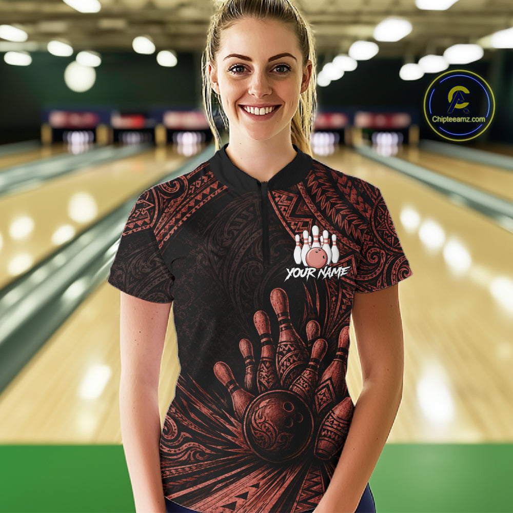 A woman standing in a bowling alley wearing a black short-sleeve bowling shirt with a red tribal hand and bowling ball graphic, and a white customizable name print on the chest.