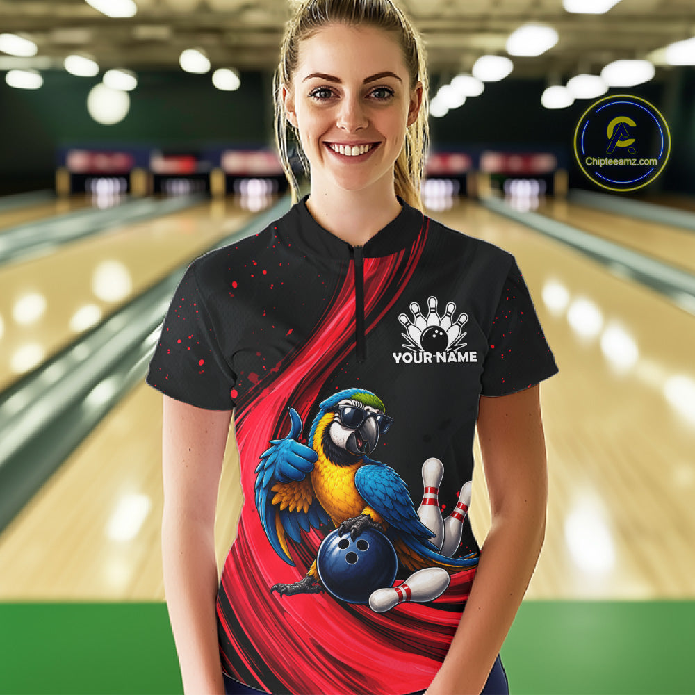 A woman wearing a black bowling-themed short-sleeve polo shirt with a red graphic swirl, a parrot illustration, bowling pins, and a customizable name print on the chest. The background shows a blurred bowling alley with lanes and pins.