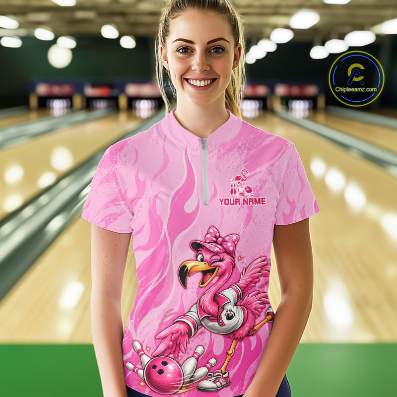 A smiling woman stands in a bowling alley wearing a pink short-sleeve zip-front shirt with a flamingo graphic and the text 'YOUR NAME'. Bowling lanes are blurred in the background, and a circular 'Cheleware' logo appears in the top right corner.