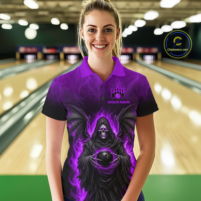 A woman stands in a bowling alley wearing a purple and black short-sleeve polo shirt with a grim reaper graphic and a bowling pin target design on the chest. The background shows blurred bowling lanes and pins.