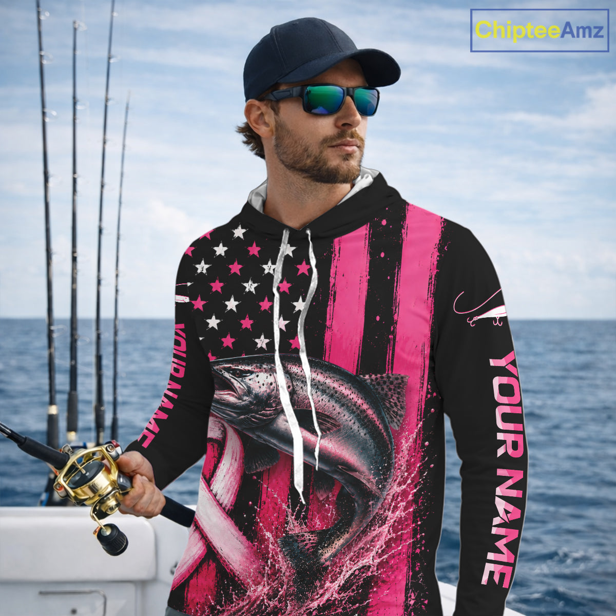 A man wearing a custom black and pink fishing hoodie with a fish graphic and 'YOUR NAME' printed on the sleeve, standing on a boat with fishing rods and the ocean in the background.