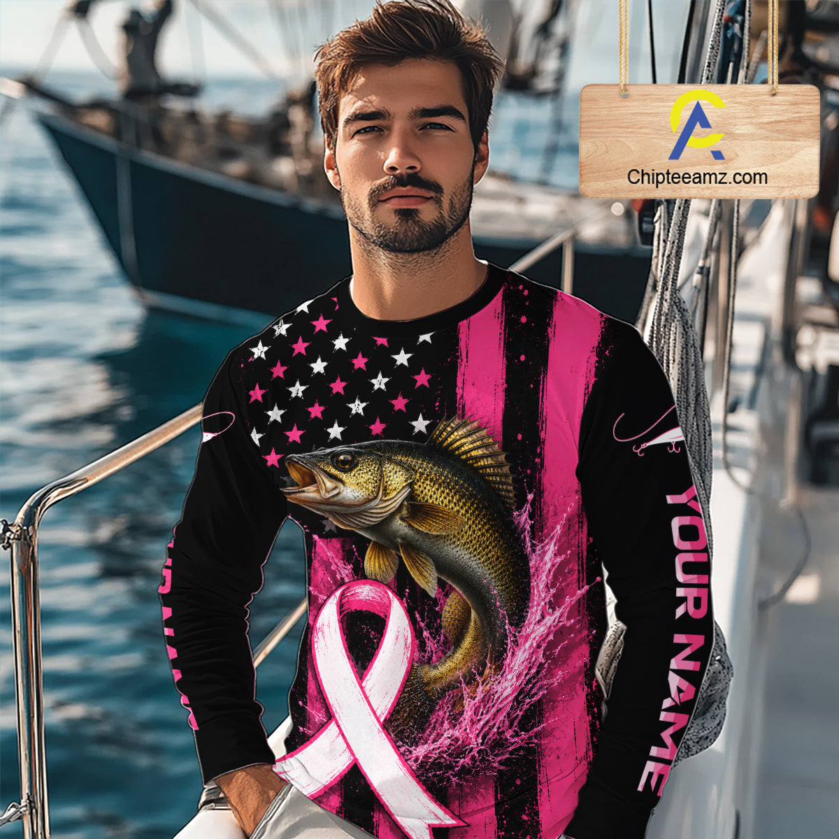  man wearing a long-sleeve black and pink graphic shirt with a large fish and pink ribbon print, standing on a boat deck with water and boats in the background. A wooden sign with the text 'Chipteeamz.