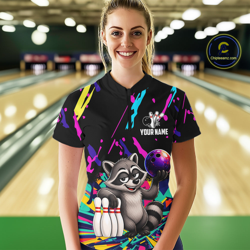 Person wearing a black bowling shirt with colorful design and raccoon graphic in a bowling alley.